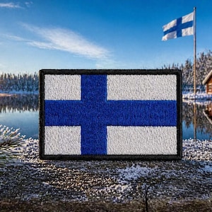 May include: Embroidered patch featuring the flag of Finland, a blue cross on a white background, set against a winter landscape with a lake, snow-covered trees, and a small wooden cabin. A Finnish flag waves in the background.