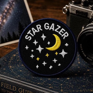 Puede incluir: Un parche bordado circular con fondo negro que presenta el texto blanco STAR GAZER en la parte superior. El diseño incluye una luna creciente amarilla y varias estrellas blancas y amarillas de cuatro puntas dispersas.