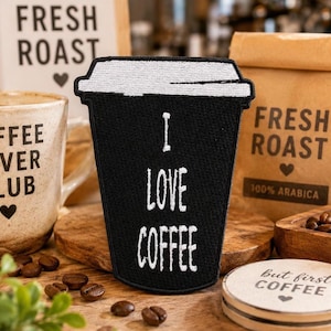 May include: A black embroidered patch shaped like a coffee cup with a white lid and the white text I LOVE COFFEE. The patch features a textured fabric surface with a stitched border, designed to resemble a disposable beverage container.