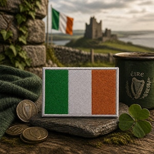 May include: A rectangular embroidered patch featuring the Irish flag with vertical stripes of green, white, and orange, finished with a light gray stitched border. The patch is displayed on a stone surface alongside a green mug and coins.