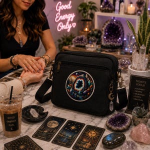May include: Black crossbody bag with a colorful geode patch. The bag has a long strap and a zipper closure. Tarot cards and crystals are on the table. The text "Good Energy Only" is in neon. A sign reads "Today's Focus".