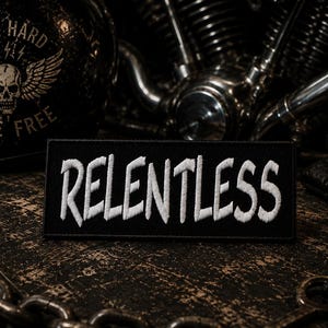 May include: A rectangular black fabric patch featuring the word RELENTLESS embroidered in white capital letters. The patch has a clean, straight-edged border and is designed for attachment to clothing or gear, showcasing a bold, high-contrast design.