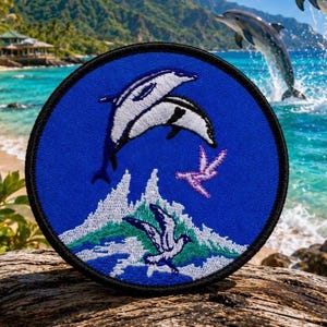 May include: A circular embroidered patch featuring a blue background with a white and black dolphin leaping above a stylized white and green wave. A small purple bird is flying near the wave, and a white bird is positioned below the wave.