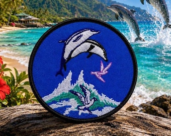 Op de afbeelding: Een ronde geborduurde patch met een blauwe achtergrond waarop een wit-zwarte dolfijn boven een gestileerde wit-groene golf springt. Een kleine paarse vogel vliegt bij de golf en een witte vogel bevindt zich onder de golf.