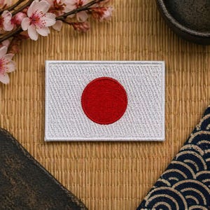 Op de afbeelding: Geborduurde patch met de vlag van Japan. De rechthoekige patch heeft een witte achtergrond met een rode cirkel in het midden. De patch is omgeven door een witte rand en is geplaatst op een natuurlijke geweven achtergrond.