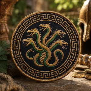 Puede incluir: Un parche bordado circular que presenta una criatura hidra de varias cabezas en hilo dorado y verde sobre un fondo negro. El diseño está encerrado por un borde dorado con patrón de greca, creando un emblema detallado con textura.