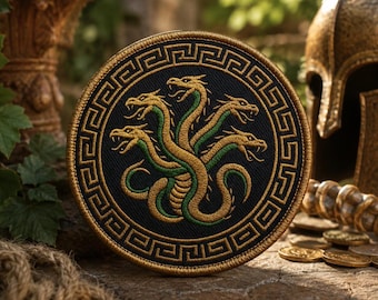 May include: A circular embroidered patch featuring a multi-headed hydra creature in gold and green thread against a black background. The design is enclosed by a gold Greek key border pattern, creating a detailed emblem with a stitched texture.