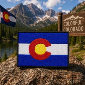 May include: A rectangular embroidered patch featuring the Colorado state flag design, which consists of three horizontal stripes in blue, white, and blue, with a large red letter C surrounding a yellow circle in the center of the patch.