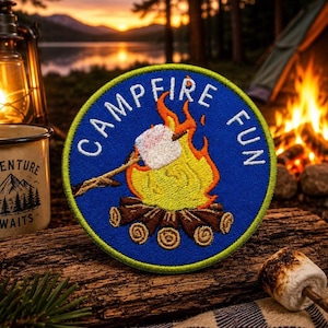 Op de afbeelding: Een ronde geborduurde patch met een blauwe achtergrond en een limoengroene rand. Het ontwerp toont een kampvuur met bruine blokken, gele en oranje vlammen en een witte marshmallow op een stokje. De tekst CAMPFIRE FUN is in het wit geborduurd.