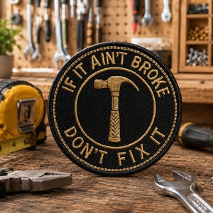以下が含まれることがあります： 「IF IT AIN'T BROKE DON'T FIX IT」のテキストとハンマーのグラフィックが入った黒と金のパッチ。メジャー、ペンチ、レンチ、ネジが木製の表面に置かれています。背景には工具と植物があります。