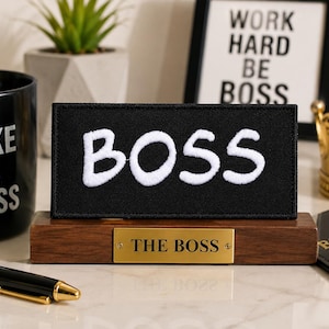Könnte beinhalten: Ein rechteckiger schwarzer Aufnäher mit dem weißen gestickten Text BOSS, montiert auf einem dunkelbraunen Holzsockel mit einer goldfarbenen Metallplakette, auf der der Text THE BOSS eingraviert ist, zentriert auf einer Fläche mit Büroartikeln.