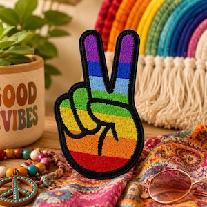 Puede incluir: Un parche de tela con forma de mano haciendo el signo de la paz, con rayas horizontales en rojo, naranja, amarillo, verde, azul y morado. El parche tiene un borde grueso bordado en negro que delinea los dedos y la palma de la mano.