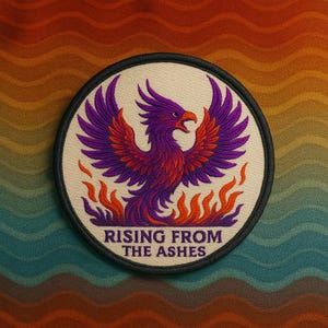 May include: Round embroidered patch featuring a purple phoenix rising from orange flames. The phoenix has outstretched wings and a red beak. The text "RISING FROM THE ASHES" is below the phoenix, with a black border.