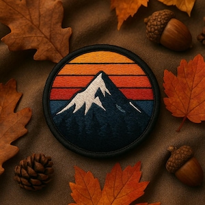 May include: A circular embroidered patch featuring a mountain scene with a sunset design. The patch has a black border, with a white mountain peak over a blue base, and orange, red, and yellow stripes above. Autumn leaves, acorns, and a pine cone surround the patch.