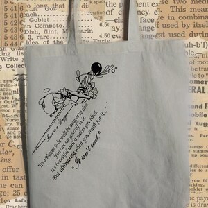 May include: A gray tote bag with a black and white floral design and the text "Love is a Dagger" and "It's a weapon to be wielded far away or up close. You can see yourself in it. It's beautiful until it makes you bleed... But ultimately when you reach for it... "It isn't real.""