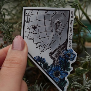 Peut inclure: Une illustration en noir et blanc d'un personnage du film d'horreur Hellraiser. Le personnage a une grille d'épines sur le visage et est entouré de fleurs bleues. Le texte "We'll tear your soul apart" est écrit sur le côté droit de l'image.