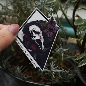 May include: A white sticker with a black and white illustration of a person wearing a ghost face mask. The sticker has the text "Do you like scary movies?" printed on it.