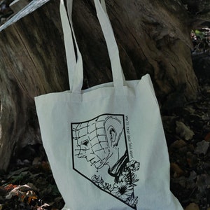 May include: A white tote bag with a black and white illustration of a character from the horror movie "Hellraiser". The character is wearing a grid-like mask and has a floral design around the head. The text "We'll tear your soul apart" is printed on the bag.