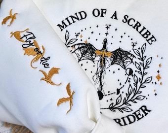 May include: White sweatshirt with embroidered design. The design includes a dragon, stars, and the text "Mind of a Scribe Rider." The sleeve features the text "Fly or Die" with gold dragon illustrations.