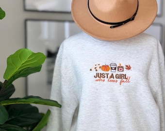 Könnte beinhalten: Ein grauer Pullover mit dem Text "JUST A GIRL who loves fall" auf der Vorderseite gestickt. Der Text ist von gestickten Bildern eines Kürbisses, einer Tasse Kaffee, einer Kerze und eines Blattes umgeben.