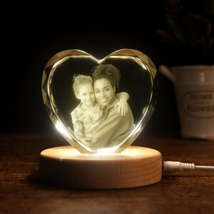 Puede incluir: Un cristal en forma de coraz&oacute;n con una foto de una madre y su hija en el interior, iluminado por una luz c&aacute;lida. El cristal est&aacute; sobre una base de madera con un cable.