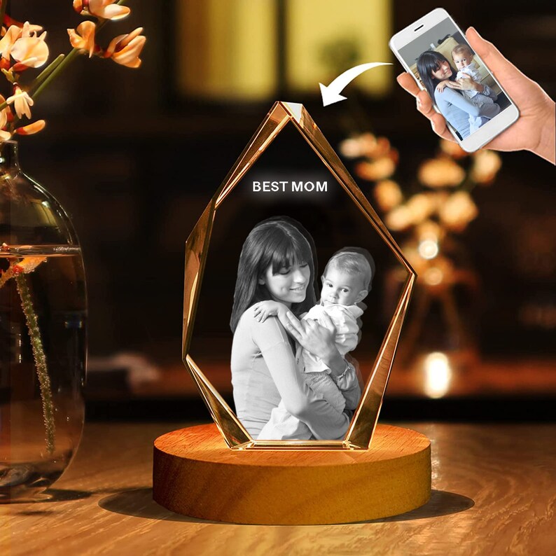 Puede incluir: Un pisapapeles de cristal con forma de diamante con una foto en blanco y negro de una madre y un ni&ntilde;o en su interior. El pisapapeles est&aacute; sobre una base de madera y tiene las palabras "BEST MOM" grabadas en la parte delantera.