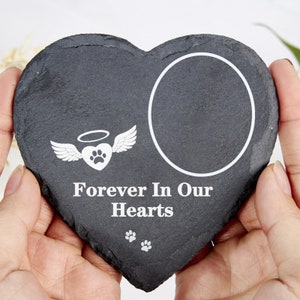 May include: A heart-shaped, dark grey slate memorial stone with a white circular photo frame. The stone features a white graphic of a heart with wings and a paw print, and the text "Forever In Our Hearts."