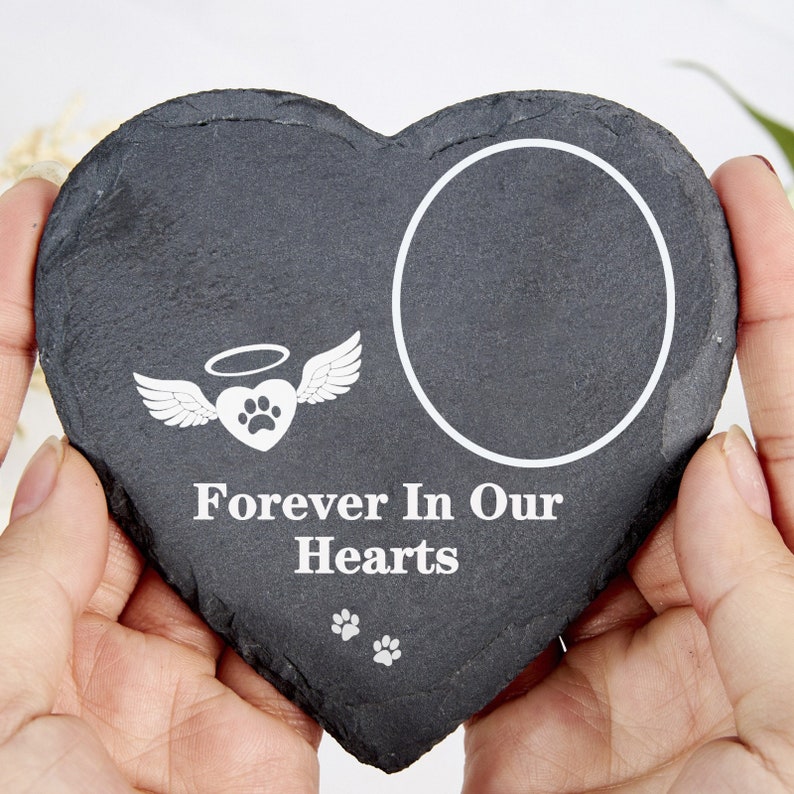 May include: A heart-shaped, dark grey slate memorial stone with a white circular photo frame. The stone features a white graphic of a heart with wings and a paw print, and the text "Forever In Our Hearts."