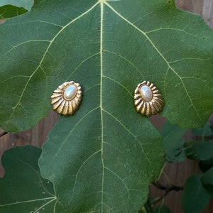 May include: A pair of gold tone earrings with a pearl center and a scalloped design. The earrings are on a green leaf.