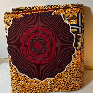 May include: A folded piece of fabric featuring a large, central, scalloped-edged circular motif in deep red and black with radiating lines. The surrounding pattern consists of vibrant orange sections with black and white speckled and geometric designs.