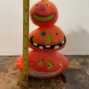 May include: A stacked Halloween candle shaped like three smiling jack-o'-lanterns. The orange candle has green, purple, and black accents. The candle measures approximately 7 inches tall.