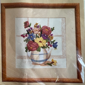 May include: A framed embroidery of a colorful floral arrangement in a white pitcher with blue stripes. The flowers are pink, yellow, purple, and blue. The embroidery is on a white background with a window pane design.