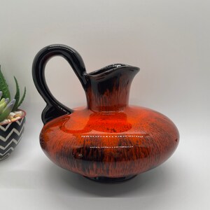 May include: A ceramic pitcher with a black handle and a brown and orange glaze. The pitcher has a rounded body and a wide mouth.