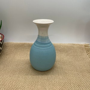May include: A small, light blue ceramic vase with a white rim. The vase has a rounded body and a narrow neck.