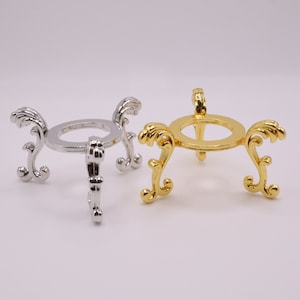 May include: Two decorative metal stands for displaying crystal balls. One stand is silver and the other is gold. Both stands have a round base and three legs with ornate designs.