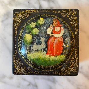 Op de afbeelding: Een zwarte houten doos met een geschilderd tafereel van een vrouw in een rode jurk en een witte hoofdbedekking die met een wit lam in een groen veld staat. De doos heeft gouden details die rond de randen zijn geschilderd.