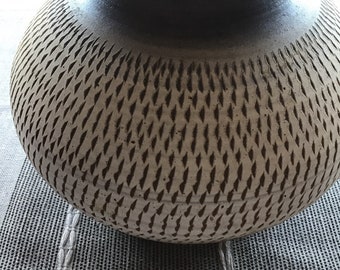Op de afbeelding: Een grote, ronde, witte keramische vaas met een donkerbruine rand en een textuurpatroon van kleine, donkerbruine lijnen. De vaas staat op een geweven tapijt.