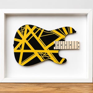 May include: A framed wall art piece featuring a stylized electric guitar body silhouette. The guitar body is black with a bold, geometric yellow striped pattern. A light-colored wooden fretboard section extends from the right side of the body.