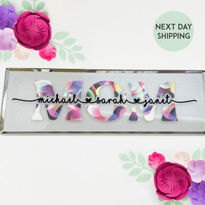 May include: A rectangular glass sign with the word "MOM" in large, colorful letters. The letters are decorated with a swirling pattern of pink, purple, and blue. Below the letters, the names "michael", "sarah", and "janet" are written in black cursive with small hearts between each name.