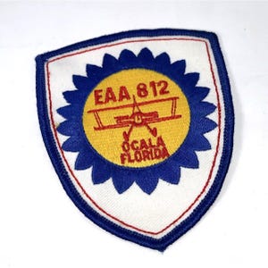 May include: Embroidered patch with a white background and a blue scalloped border. The patch features a yellow circle with a red biplane and the text "EAA 812 OCALA FLORIDA". The patch has a red border.
