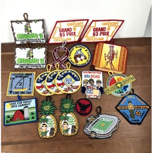 May include: A collection of colorful Cub Scout patches with various designs and text. Patches include text such as "Grand Prix," "Extravaganza," and "Lake Cub Family Campout." Some patches feature images of pineapples, a stadium, and a sailboat.