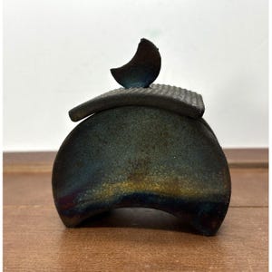May include: A handcrafted ceramic container with a lid and a crescent moon-shaped accent. The container has a dark, textured surface with hints of blue, green, and gold. The lid is rectangular with a ridged texture.