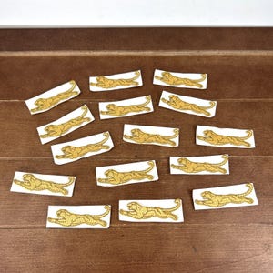 May include: A collection of gold-colored embroidered patches, each featuring a running tiger design. The patches are rectangular with a white background and are arranged on a brown wooden surface. The tiger's body is detailed with gold thread.