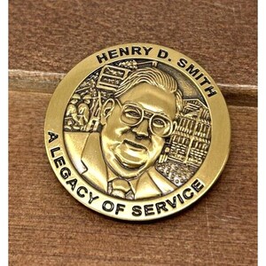 May include: A gold-colored commemorative coin featuring a portrait of Henry D. Smith, with the text "HENRY D. SMITH" and "A LEGACY OF SERVICE" encircling the image. The coin has a detailed design with architectural elements.
