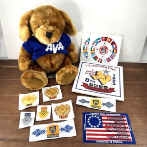 May include: A plush brown dog wearing a blue shirt with "AVA" in white letters. Assorted patches and a sign with text including "VOLKSSPORTING" and "Liberty Bell Wanderers" are also present. The patches feature numbers like 650, 700, and 550.