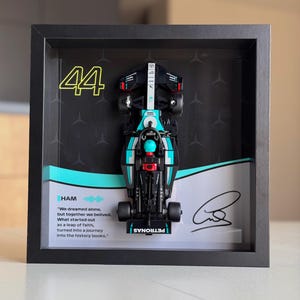 Puede incluir: Una foto enmarcada en blanco y negro de un coche de carreras de Fórmula 1 en miniatura con el número 44 en el lateral. El coche está en un marco negro con fondo blanco. La cita "We dreamed alone, but together we believed. What started out as a leap of faith, turned into a journey into the history books." está impresa debajo del coche. La cita se atribuye a HAM.