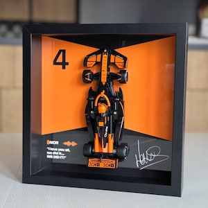 Peut inclure: Une voiture de course de Formule 1 noire et orange encadrée avec le numéro 4 sur le côté. La voiture est signée par le pilote, avec le texte "NOR "I love you all, we did it... WE DID IT!" en dessous de la voiture.