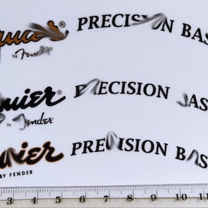 Może przedstawiać: Trzy różne wersje naklejki Squier by Fender Precision Bass. Pierwsza naklejka jest złota z czarnym tekstem. Druga naklejka jest czarna z białym tekstem. Trzecia naklejka jest czarna ze złotym tekstem. Wszystkie trzy naklejki mają tekst "Squier by Fender" i "Precision Bass".
