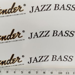 以下が含まれることがあります： 白い背景に3つのFender Jazz Bassデカール。 Fenderのロゴは、茶色と金のグラデーションの様式化されたスクリプトです。 テキスト「JAZZ BASS」と「OFFSET Contour Body」は黒です。 下部に定規があります。