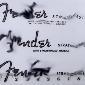 Op de afbeelding: Drie Fender Stratocaster logo's met tekst, waaronder "WITH SYNCHRONIZED TREMOLO" en patentnummers. Een liniaal met millimeter- en centimeter markeringen staat onderaan. De logo's zijn in een gestileerd lettertype.